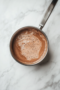 In this top-down photo, the hot chocolate mixture simmers gently in a silver saucepan for 5–7 minutes. A light steam rises from the surface, which is now a velvety deep brown. All the added spices—vanilla, cinnamon, chili, nutmeg, and cloves—infuse the drink with a warm, fragrant aroma. The scene conveys comfort and care in preparation.