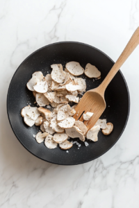 Top-down view of sliced mushrooms sautéing in a black skillet with olive oil, salt, and freshly cracked black pepper on a white marble countertop. The mushrooms are beginning to brown and release their juices, filling the kitchen with a savory aroma. This step builds the earthy base of the pasta sauce.