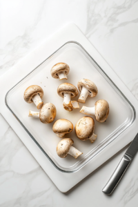 Top-down image showing the process of preparing mushrooms by slicing and separating stems from caps. A white cutting board holds fresh mushrooms while a knife gently removes the stems. The mushroom caps are clean and ready to be stuffed. This image highlights the essential first step of prepping mushrooms before cooking.