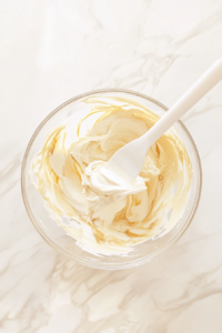 Confectioners' sugar is gradually being added to the cream cheese mixture while an electric mixer blends everything together. The mixture becomes light, smooth, and fluffy, forming the perfect base for the dessert.