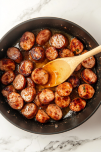 Smoked sausage slices are sizzling in melted butter inside a large black pot over a white marble countertop. The sausage pieces are turning golden brown, releasing rich aromas. A wooden spoon stirs the slices, ensuring even cooking. The butter glistens on the surface, adding flavor and depth to the dish.