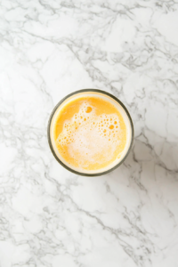 Top-down view of a frothy yellow mixture inside a glass blender placed on a white marble kitchen countertop. The blend of coconut milk, pineapple juice, rum, sugar, and ice is now fully mixed and smooth. The thick, airy texture is visible, promising a rich and refreshing tropical beverage ready for serving.