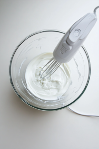 A large glass bowl on a clean white marble countertop holds softened cream cheese being whipped with a white hand mixer. The cream cheese is visibly smooth and creamy. An empty cream cheese wrapper lies nearby. This step focuses on preparing the rich filling that will top the cake mix crust in the next stage.