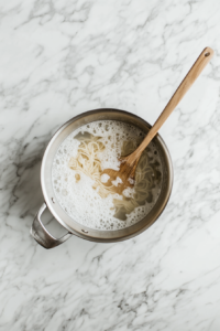 A top-down shot of spaghetti noodles boiling in a large silver pot filled with water on a white marble countertop. The noodles are halfway cooked and surrounded by bubbling water. This step is essential for softening the pasta before combining it with the rich flavors of teriyaki sauce in the next steps.