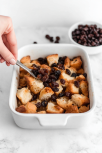 Top-down view of raisins being sprinkled over torn bread pieces arranged in a buttered white ceramic baking dish. The raisins add sweetness and chewy texture to the pudding. A small white bowl with extra raisins sits nearby. All items are neatly placed on a white marble countertop, creating a clean baking environment.