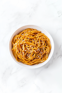 A delicious bowl of stir-fried soy garlic noodles served in a clean white ceramic bowl placed on a white marble countertop. The noodles are golden brown and glossy, coated in a savory soy and teriyaki sauce, and garnished with white sesame seeds. Bits of sautéed garlic, ginger, and onions are visible, making it look rich and flavorful. The minimal, bright background keeps the focus on the warm and comforting dish, ready to be served hot.