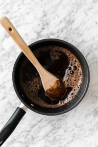 The chocolate mixture is bubbling vigorously in a black saucepan as it reaches a rolling boil. A wooden spoon is stirring constantly to prevent burning and ensure even mixing. The white marble countertop underneath highlights the process. This boiling step is key to properly melting sugar and developing the cookie texture.