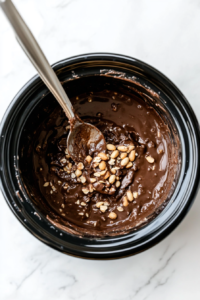 A silver spoon stirs the fully melted mixture of white chocolate, semisweet chocolate, German sweet chocolate, and roasted peanuts inside a black crock pot. The texture is creamy, rich, and smooth, with every peanut evenly coated in chocolate. This is a crucial step that ensures the consistency is perfect for scooping.