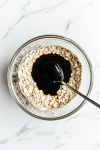 Top-down view of the melted syrup mixture being stirred into the dry oat and flour blend inside a large clear glass mixing bowl. The mixing is halfway through, creating a thick, sticky dough. The blending of wet and dry ensures every ingredient is coated with the treacly sweetness, setting the base texture for baking.