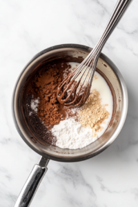 Top-down view of vanilla extract, cinnamon, chili powder, nutmeg, and ground cloves being stirred into the warm cocoa mixture using a stainless steel whisk. The mixture is turning darker and more aromatic inside the silver saucepan, resting on a clean white marble surface. Measuring spoons with spice traces are nearby, showing active preparation of this flavorful drink.