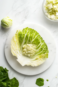 Top-down view of a cabbage leaf laid flat on a cutting board placed over a white marble countertop. Two tablespoons of the filling are placed in the center, ready to be rolled. A spoon and a few rolled pieces are visible. This step captures the art of wrapping the filling tightly into soft cabbage leaves to form compact rolls.
