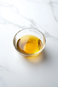 A simple dressing made of sunflower oil, vinegar, and salt is being whisked in a small clear glass bowl. The bowl is placed on a pristine white marble surface, and measuring spoons are nearby. This dressing brings a perfect balance of tang and smoothness, ready to be poured over the crisp, colorful cabbage and cucumber salad.