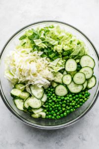 Top-down image showing bright green sweet peas after being thawed in cold water and drained, ready to be added to the glass bowl with cabbage, cucumber, and green onions. The peas bring a touch of natural sweetness and texture to the salad, and the white marble countertop adds a clean aesthetic.