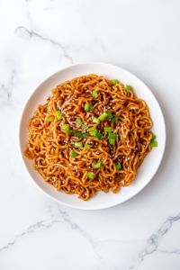 A top-down view of delicious teriyaki noodles served on a white ceramic plate, garnished with chopped green onions and sesame seeds. The noodles are perfectly coated in a glossy teriyaki sauce and look warm, comforting, and flavorful. This simple and quick meal is great for busy weeknights or casual lunches.