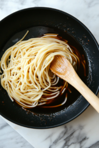 A top-down shot showing teriyaki sauce being poured over spaghetti noodles in a black nonstick pan. The sauce begins to coat the noodles with a dark, rich glaze. This step brings deep umami flavor to the dish, turning plain noodles into a savory and satisfying Asian-style stir fry.