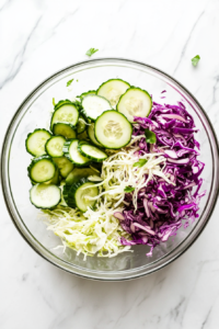 Thin cucumber slices are added into a large clear glass bowl already filled with shredded white and red cabbage. The mixing bowl rests on a white marble countertop, and a cutting board with remaining cucumber slices is nearby. The refreshing green cucumber contrasts beautifully with the bold cabbage colors, enhancing the salad’s fresh and crunchy texture.