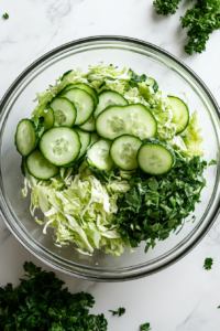 Top-down image showing thin half-circle slices of an English cucumber being prepared on a wooden cutting board beside a sharp knife. The slices are added to a glass bowl containing previously shredded cabbage, resting on a white marble countertop. The focus is on the crisp freshness of the cucumber, adding crunch to the salad.