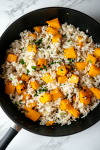 Arborio rice is stirred into the pumpkin, onion, and garlic mixture in the same black skillet. The grains are beginning to toast slightly, turning translucent, which is essential for developing the risotto’s creamy texture. The simple setup includes the remaining ingredients in glass bowls, ready to be incorporated, all placed on a white marble countertop.