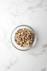 A clear glass bowl filled with freshly cleaned and thoroughly dried pumpkin seeds sits on a white marble countertop. The seeds are pale in color and spread evenly in the bowl, with a soft white cloth next to it, suggesting gentle drying. The clean, minimalistic setup emphasizes this crucial preparation step before mixing the seeds with oil and honey.