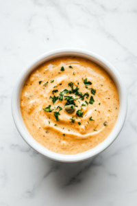 A top-down view of a white ceramic bowl filled with rich, creamy remoulade sauce. The sauce is light orange in color with visible flecks of chopped herbs and spices, giving it a textured appearance. The bowl is placed on a simple white marble countertop with no extra elements, focusing entirely on the freshness and creamy consistency of the homemade sauce perfect for serving with seafood or fried snacks.