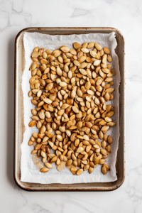 Pumpkin seeds coated with oil and honey are evenly spread on a parchment-lined silver baking sheet, ready for roasting. The seeds glisten under soft lighting, showing an even distribution. The baking sheet is placed over a white marble countertop, reinforcing the clean and simple kitchen aesthetic. This image visually prepares viewers for the roasting step.