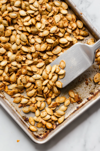 This image captures a hand stirring golden brown pumpkin seeds halfway through roasting on a baking tray lined with parchment. A silver spatula gently moves the seeds, ensuring even roasting. The seeds have started to caramelize, showing early signs of a crispy finish. The baking tray rests on a white marble countertop, keeping the visual setting minimal and tidy.