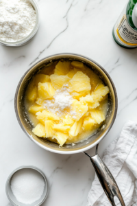 Top-down image of the pineapple topping cooking in a small nonstick saucepan. The mixture includes crushed pineapple, lime juice, sugar, cornstarch, and optional coconut rum, now thickened into a glossy, fruity layer. A small whisk lies nearby and measuring spoons with lime juice and sugar sit beside the pan on a white marble surface.