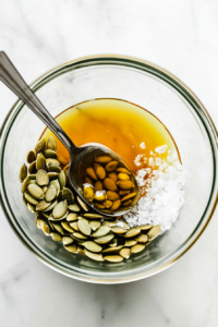 This top-down image shows a glass mixing bowl containing raw pumpkin seeds combined with pumpkin seed oil, runny honey, and a pinch of flaky sea salt. A silver spoon stirs the ingredients, evenly coating the seeds. The glossy shine from the honey and oil is visible. The clean white marble background ensures full attention on the mixing process.