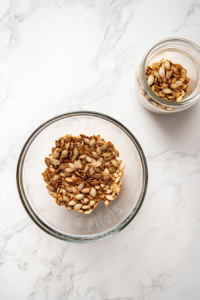 A glass storage jar filled with crispy roasted pumpkin seeds sits next to a clear bowl of the remaining seeds on a white marble countertop. The seeds appear crunchy and golden, ready to be enjoyed or stored. This final image completes the process, showing the recipe’s long-lasting appeal and simple storage in a clean, attractive presentation.