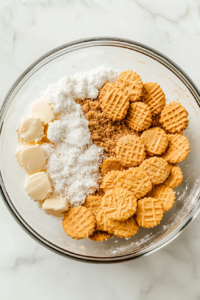 A top-down image showing the crushed vanilla wafers, unsweetened coconut flakes, light brown sugar, kosher salt, and melted butter combined in a clear glass bowl. The golden crumbly mixture has been pressed into a 9-inch pie pan, ready to be baked. The preparation is happening over a clean white marble countertop, capturing the crust before baking.