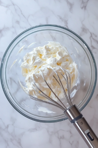 Top-down image of freshly whipped cream in a glass mixing bowl, beaten to soft peaks using a hand mixer. The ingredients—heavy cream, sugar, vanilla extract, and optional coconut cream—are seen around the bowl. The setup is clean and minimalistic, capturing the fluffy texture of the cream over a white marble countertop.