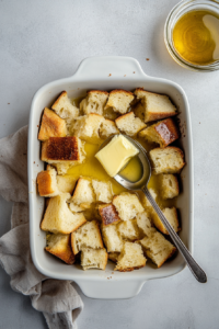 Top-down view of a white ceramic baking dish filled with torn pieces of day-old bread, as melted unsalted butter is being drizzled over it using a silver spoon. The bread pieces are irregularly shaped for a rustic texture. The setup is placed on a clean white marble countertop to highlight baking preparation.