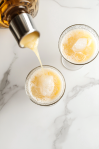 Top-down shot of a tropical drink being poured from a glass blender into two cocktail glasses on a clean white marble countertop. The creamy yellow drink flows smoothly, containing blended coconut milk, pineapple juice, rum, sugar, and ice. This image captures the moment just before enjoying the refreshing island-inspired beverage.