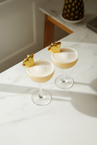 Top-down view of two tropical cocktails made with coconut milk, pineapple juice, rum, sugar, and ice, served in clear cocktail glasses. The drinks are creamy yellow, frothy, and garnished with fresh pineapple slices and cherries. They sit on a white marble countertop, creating a clean, refreshing, and inviting tropical presentation perfect for summer.