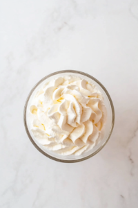 A clear glass bowl filled with heavy cream is whipped to stiff peaks using an electric hand mixer, sitting on a clean white marble countertop. The cream appears light and airy, ready to be folded into the chocolate mixture later. This crucial step ensures the final cheesecake texture is light and mousse-like.