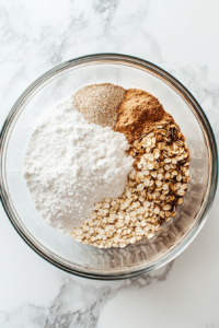Top-down view of a large glass mixing bowl filled with whole wheat flour, rolled oats, white sugar, baking powder, and baking soda. These dry ingredients are being whisked together on a clean white marble countertop. A stainless steel whisk and measuring spoons are visible, emphasizing the clean and organized setup.