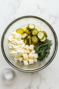 In a large clear glass mixing bowl placed on a white marble countertop, mayonnaise, sugar, sweet pickle relish, dill pickle relish, white vinegar, yellow mustard, and vegetable oil are being whisked together to create a smooth and creamy coleslaw dressing. The image captures the beginning of the flavor-building process for the coleslaw.