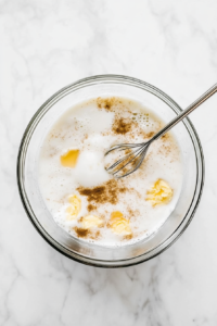 Top-down view of a large glass mixing bowl placed on a white marble countertop, filled with milk, beaten eggs, white sugar, cinnamon, and vanilla extract being whisked together with a silver whisk. This mixture forms the flavorful custard base for the bread pudding, ensuring a rich and creamy texture when baked.