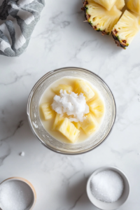 Top-down view of rum and white sugar being added to a blender already containing coconut milk and pineapple juice. The clear glass blender rests on a clean white marble surface, with a sugar bowl and rum bottle nearby. The image captures the careful addition of ingredients needed to enhance the flavor of the tropical cocktail.