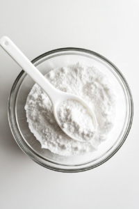 A top-down view of a clear glass bowl filled with white sugar, cornstarch, and ground cinnamon being gently stirred with a white silicone spoon. All ingredients are precisely measured and laid out on a white marble countertop. This step brings together the dry components that will add sweetness, thickness, and spice to the pudding.