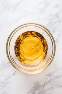 A spoonful of white vinegar is being stirred into the thick, flavorful sauce mixture. The image showcases the clear vinegar blending into the mix, adding tanginess. A small bottle of vinegar and a measuring spoon are placed next to the bowl, all arranged neatly on the pristine white marble countertop.