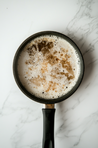White wine has just been added to the sizzling mixture of mushrooms, garlic, and butter inside a black skillet on a white marble countertop. The wine is bubbling slightly, releasing a fragrant steam as it reduces. This step enhances the flavor and brings a restaurant-quality depth to the mushroom dish.